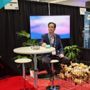 RiskWorld 2025 exhibit hall: Bill Nagle sits at a high-top table branded “Cloud Claims,” beside a wire basket overflowing with “This Is Fine” plush dogs and APPTech materials.