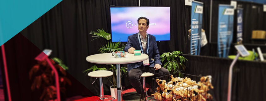 RiskWorld 2025 exhibit hall: Bill Nagle sits at a high-top table branded “Cloud Claims,” beside a wire basket overflowing with “This Is Fine” plush dogs and APPTech materials.